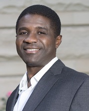 Firmin Charlot is smiling and is wearing a white shirt and a grey jacket.